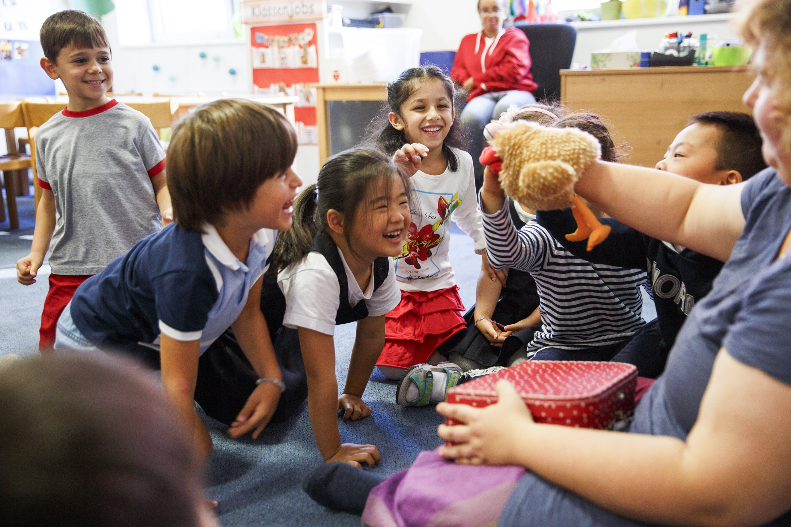 Eine Gruppe lächelnder Kinder sitzt um ihre Erzieherin herum, welche eine Puppe in der Hand hält und ihnen etwas erzählt.