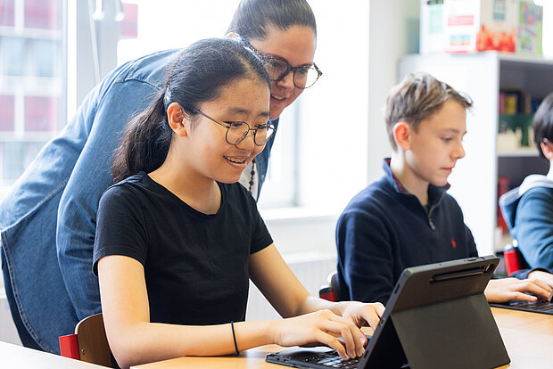 Eine Schülerin schreibt etwas auf einem IPad, während eine Lehrerin ihr über die Schulter guckt im Phorms Gymnasium Hamburg