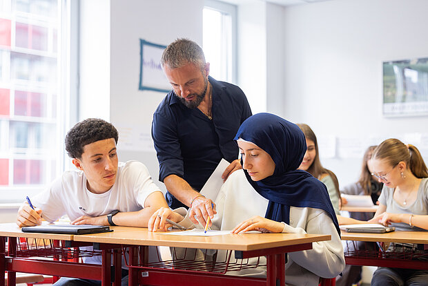 Schüler sitzen im Unterricht und der Lehrer erklärt ihnen etwas auf ihrem Arbeitsblatt im Phorms Gymnasium Hamburg