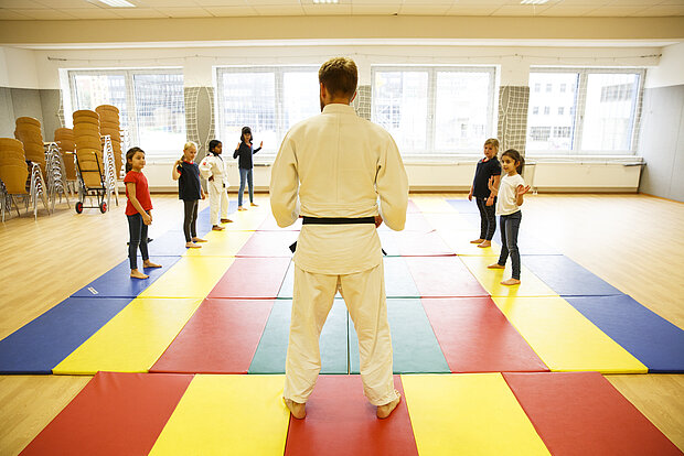 Sechs Kinder stehen im Sportraum mit einem Sportlehrer, der einen Kampfsport Anzug an hat in der Phorms Grundschule Hamburg