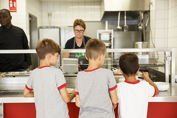 Drei Kinder stehen in der Kantine an der Theke und nehmen sich ihr Mittagessen an der Phorms Grundschule Hamburg