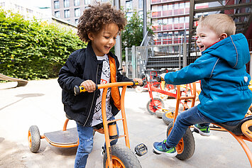 Phorms Hamburg Kita Kinder auf Dreirad Zwei lächelnde Kindergarten-Kinder auf Dreirädern spielen auf dem Schulhof auf Dreirädern