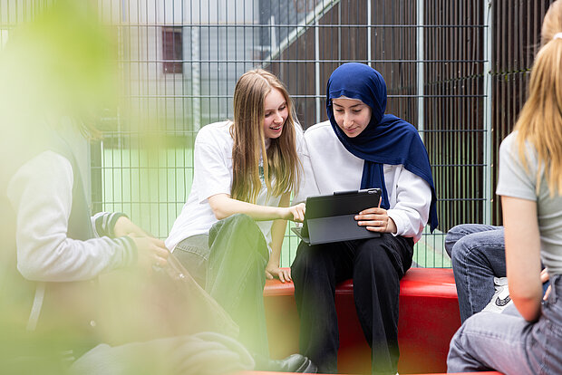 Zwei Schülerinnen sitzen gemeinsam in der Hofpause und gucken auf ein Ipad auf dem Schulhof des Phorms Gymnasiums Hamburg