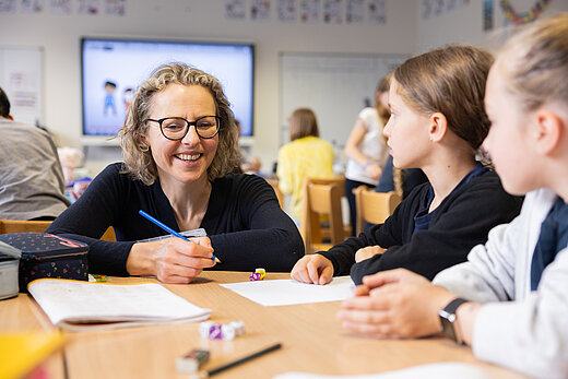 Zwei Kinder sitzen im Klassenraum mit Lehrerin, die ihnen etwas erklärt.