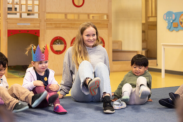 Eine lächelnde Erzieherin sitzt mit lächelnden Kindern im Morgenkreis während einer Aktivität in der Kita Phorms Hamburg