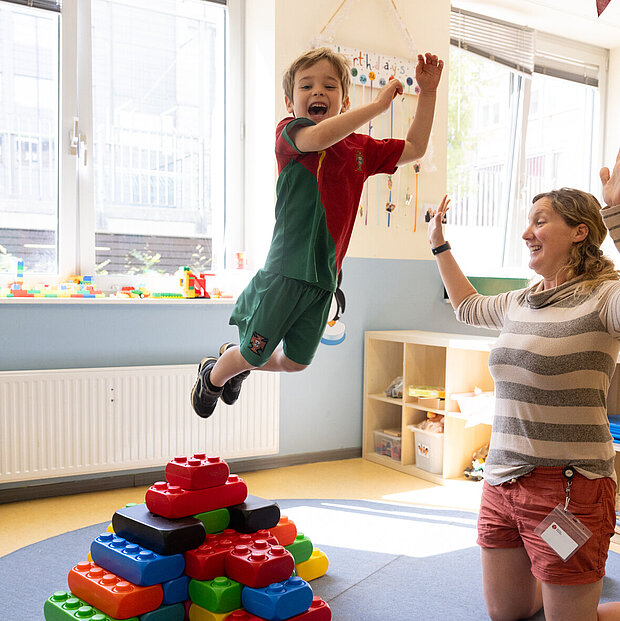 Ein Kind springt von gestapelten Blöcken unter Aufsicht der Erzieherin in der Phorms Kita Hamburg